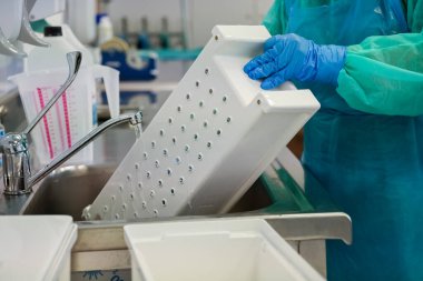 Sağlık çalışanlarının eldivenli ellerine yakın çekim bir laboratuarda akan suyun altındaki tıbbi alet tepsisini temizleme, hastanın güvenliği için yoğun sterilizasyonu vurguluyor