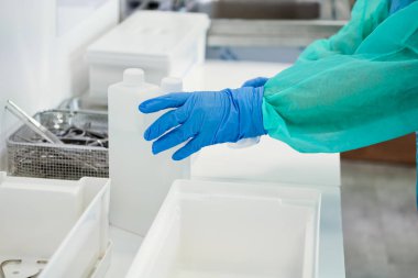 Tıp işçisinin eldivenli ve koruyucu kolunun yakın çekimi steril klinik ortamında cerrahi aletlerin yanında dezenfektan solüsyonu şişeleriyle ilgilenmek.