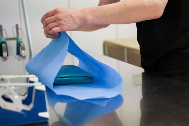 Tıbbi tesislerde hijyenin korunması için gerekli olan koruyucu steril bir çarşaf ile mavi bir tıbbi malzemeyi dikkatlice saran bir kişinin ellerine yakın çekim.