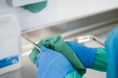 Sağlık görevlisinin eldivenli elleri tek kullanımlık yeşil bir bezle ameliyat aletini temizliyor. Sterilizasyon ve hasta bakımı için hayati önem taşıyor.