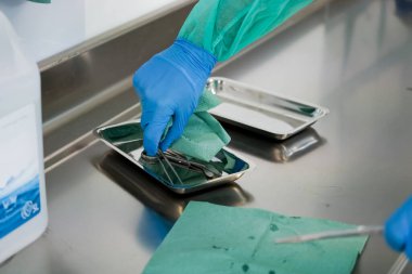 Mavi eldivenli, yeşil elbiseli, paslanmaz çelik tezgahta sterilize edilmiş cerrahi aletleri dikkatlice hazırlayan bir sağlık görevlisinin yakın çekimi hasta bakımı için katı hijyen protokollerini vurguluyor.