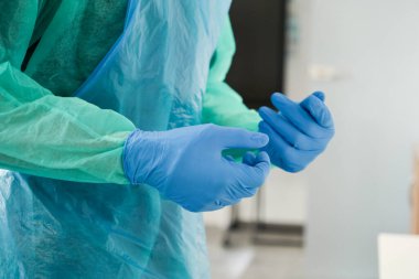 Koruyucu yeşil ve mavi tıbbi giysiler içinde mavi tek kullanımlık eldivenler takan kişi, sağlık hizmetlerinde hayati güvenlik ve hijyeni vurguluyor.