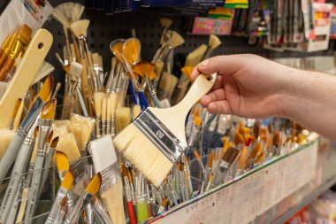 Geniş, düz bir fırçayı tutan bir elin yakın çekimi, mağazadaki çeşitli sanat fırçaları ile çevrili, sanatsal seçimi ve yaratıcı hobi keşiflerini sembolize eden