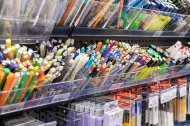 Çeşitli kalemler ve renkli kalemler de dahil olmak üzere çeşitli yazma araçları bir mağaza rafında sergilenmekte, yaratıcılığı, eğitimi ve sanat malzemeleri alışverişini sembolize etmektedir.