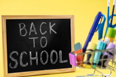 Tebeşirle okula dönüş başlıklı karatahta, canlı okul malzemeleriyle dolu minyatür bir alışveriş arabasının yanında, akademik hazırlık ve yeni öğrenmeyi vurguluyor