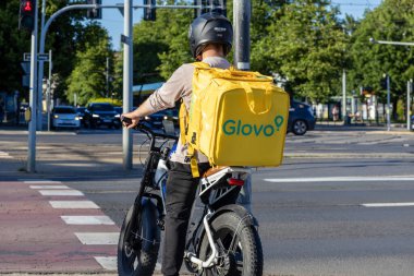 Poznan, Polonya - 30 Haziran 2025: Yoğun şehir yaya geçidinde bekleyen sarı termal çantalı bir e-bisikletli Glovo kuryesi