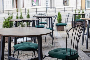 Boş ahşap masalar ve açık bir kafede konforlu yeşil yastıklı siyah metal sandalyeler, şehir konukseverliğini ve huzurlu bir atmosferi ön plana çıkarıyor.