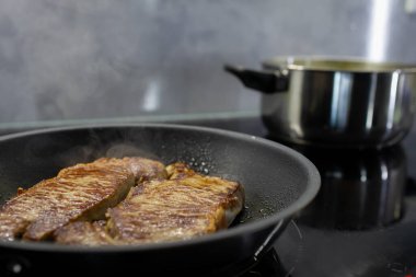 Yağda kavurulan iki kurutulmuş bifteğin yakın çekimi, buhar yükseliyor, modern bir mutfakta sade siyah bir indüksiyon fırınında.