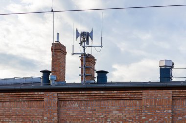 Geleneksel tuğla bina çatısı üzerindeki hava saldırısı uyarı sireninin görüntüsü, kamu alarm sistemlerini, afet hazırlığını ve kentsel güvenlik tedbirlerini temsil ediyor