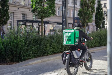 Poznan, Polonya - 16 Temmuz 2025: Yeşil çantalı bir bisikletle Uber Eats teslimatçısı, şehir caddesinde bisiklet sürüyor, modern fast food dağıtımını vurguluyor