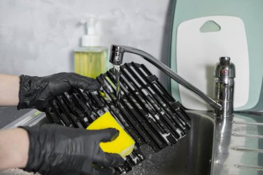 Koruyucu siyah eldivenlerle yakın plan çekimde barbekü ızgarasını parlak sarı bir süngerle güzelce fırçalarken mutfak hijyeninin altını çiziyorum.