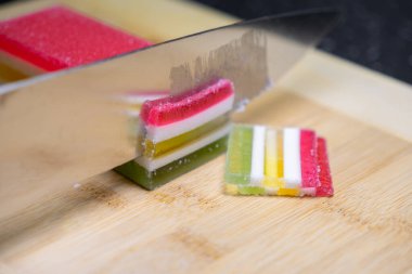 Şefin bıçağını gösteren yakın çekim sahnesinde çok katmanlı bir jöle bloğundan ayrı ayrı parçalar kesiliyor, tatlı tadı ve renkli sunumunu vurguluyor.