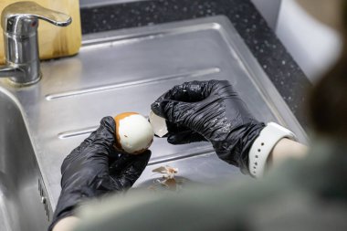 Ellerin siyah tek kullanımlık eldiven giydiği, haşlanmış yumurtayı soyarken, modern bir mutfak ortamında gıda güvenliği ve hijyeni ön plana çıkardığı yakın plan görüntüler.