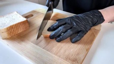 Siyah koruyucu eldivenler giyen bir kişi, taze beyaz sandviç ekmeğini bambu bir tahtanın üzerinde kesin bir şekilde dilimler, dikkatli yemek kullanımı sağlar ve profesyonel mutfak hijyenini gösterir.