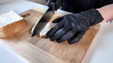 Bir insanın ellerini siyah koruyucu eldivenlerle yakından görmek keskin bir bıçakla temiz tahta bir tahtanın üzerinde yemek pişirmek, mutfak hijyeni ve gıda güvenliğini arttırmak için yumuşak beyaz ekmek hazırlamak.
