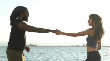 A young African American and Asian couple dance joyfully by the pier at sunset, silhouetted against golden skies.