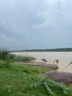 Kahverengi suyu izleyiciye doğru akan geniş bir nehir. Nehrin kıyıları yeşil bitki örtüsüyle kaplı ve kıyıda büyük silindirik nesneler var, muhtemelen sel engelleri..