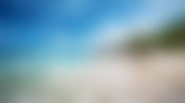 Yumuşak bir odak görüntüsü, canlı mavi gökyüzü, kumlu sahil ve yeşil yeşillik ipuçlarıyla tropikal bir plajın özünü yakalar..