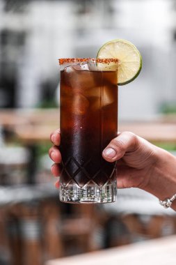 Açık terasta limonla süslenmiş Küba Libre kokteylini tutan bir el ferahlatıcı içeceği ve canlı sunumunu sergiliyor.