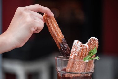 Zengin çikolata sosuna batırılmış Meksika churros 'u tutan bir el, Churros' un sıcak, altın dokusunu ve lezzetli çikolata kaplamasını sergiliyor. Yemek ve tatlı fotoğrafçılığı teması için ideal