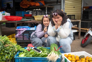 Saçları bukleli iki Vietnamlı genç kız Mong Cai, Quang Ninh, Vietnam 'da sebze ve portakal satıyorlar..