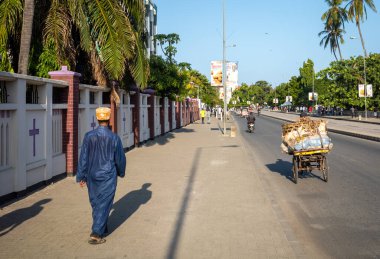 A muslim man wearing a traditional kanzu long tunic and kofia cap walks past the Azania Front Cathedral in Dar es Salaam, Tanzania. clipart