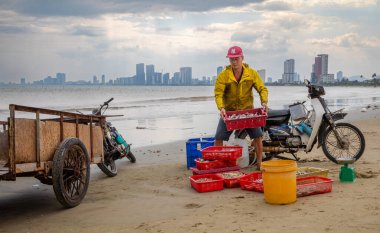 Bir balıkçı, Son Tra, Danang, Vietnam 'da My Khe plajında paslı bir scooter' ın yanında balık ve deniz ürünleri tepsisi taşır..