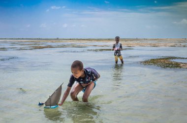 Two young boys play with toy sailing dhows made from plastic sandals on Jambiani beach, Zanzibar, Tanzania clipart