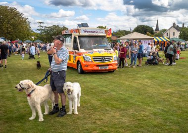 Elinde iki beyaz köpek tutan adam Wisborough Green Village Fete, West Sussex, İngiltere 'de bir dondurma kamyonunun yanında dondurma yiyor.