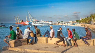 Bir grup erkek ve çocuk, Taş Şehir, Zanzibar, Tanzanya 'daki feribot limanının yanındaki deniz duvarında oturuyorlar.