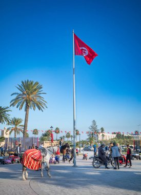 A horse waits for tourists as people meet and talk next to a Tunisian flag in Bourguiba Square, Monastir, Tunisia. clipart