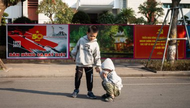 Küçük bir çocuk, Vietnam 'ın başkentinin merkezindeki Hanoi Halk Komitesi binasının önündeki Komünist Parti propaganda posterlerinin yanında küçük kardeşine yardım ediyor..