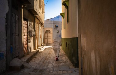 An elderly Tunisian woman dressed in a traditional foutah walks down a narrow lane inside the ancient medina of Kairouan in Tunisia. Kairouan is the 4th holiest city in Islam and is a UNESCO World Heritage Site. clipart