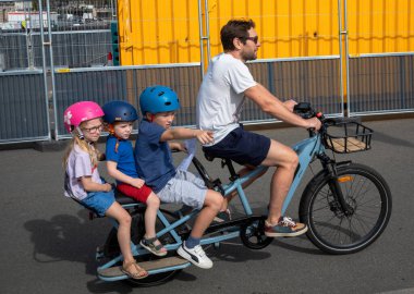 Bir çocuk, Fransa 'nın başkenti Paris' te üç küçük çocuğu sırtında taşıyan elektrikli bisiklete binen bir babayı işaret ediyor. Motosiklet bir Decathlon ELOPS BTWIN elektrikli arka yükleme uzun kuyruklu kargo bisikleti.