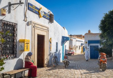 Kairouan, Tunus 'taki postanenin bulunduğu antik binanın dışında bir kadın oturuyor. Kairouan İslam 'ın dördüncü en kutsal şehridir ve UNESCO' nun Dünya Mirasları Alanıdır..