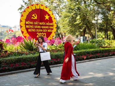 Geleneksel ao dai şapkalı bir kadın akıllı telefonuna bakar. Pahalı giyinmiş bir kadın ters yönde yürür. Orta Hanoi, Vietnam 'da komünist propaganda posterinin yanından geçer.