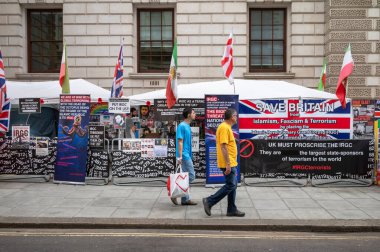 London / UK - Jul 11 2024: Two men walk past posters and slogans at the anti-Iranian government protest camp outside the Foreign, Commonwealth and Development Office in King Charles St, London. clipart