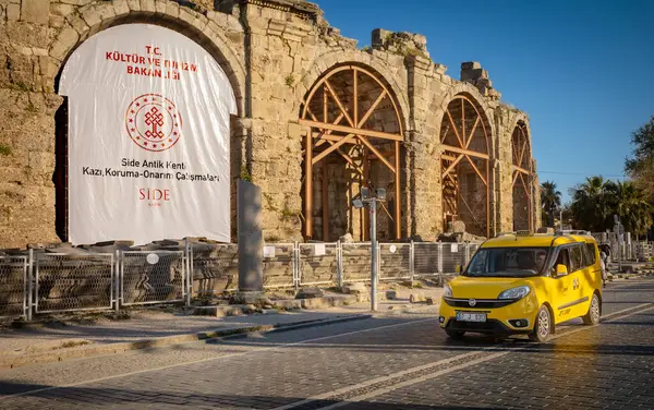 Antalya ilinin Side ilçesindeki antik Roma kentinde bulunan amfitiyatronun kalıntılarından sarı bir taksi geçiyor (Türkiye))