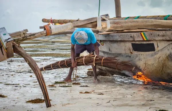 stock image A fisherman uses burning palm fronds to char and waterproof the hull of his traditional wooden dhow boat, Jambiani, Zanzibar, Tanzania