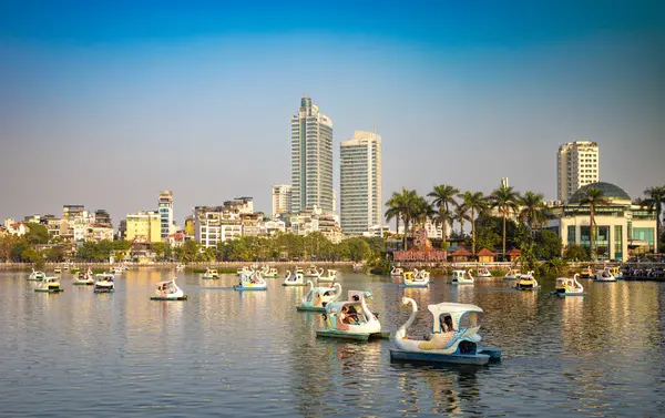 İnsanlar Truc Bach Gölü 'nde Vietnam Elektrik Karargahı veya EVN İkiz Kuleleri ile birlikte kuğu şeklinde pedallı botlara biniyorlar.