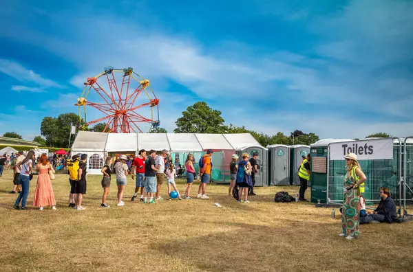 stock image Guildford / UK - Jun 29 2024: People queue for the toilets near a ferris wheel at Guilfest music festival, Guildford, Surrey, UK.