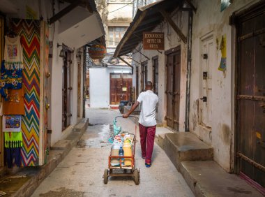 Bir çay ve içki satıcısı arabasını Stone Town, Zanzibar, Tanzanya 'da bir sokak boyunca dükkanların önünden geçiriyor..