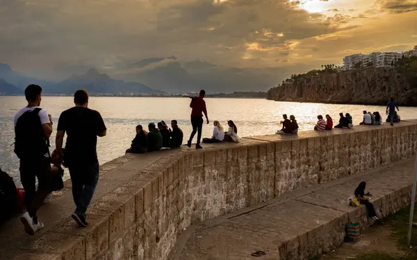 stock image People sitting and walking on the harbour wall at dusk in Antalya, Antalya Province, Turkey (Turkiye), with the sky turning orange during sunset and the Taurus Mountains (Toros Mountains) visible in the distance. The calm and peaceful scene captures 