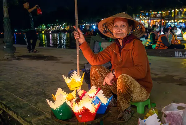 Konik şapkalı yaşlı Vietnamlı bir kadın Hoi An, Vietnam 'daki rıhtımda yanan mumlarla yüzen fenerler satıyor..