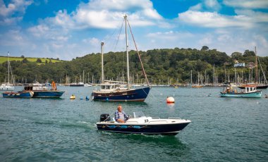 Dart Estuary, Dartmouth, Devon, İngiltere 'de demirlemiş teknelerin yanından geçen bir adamla birlikte motorlu küçük bir tekne.