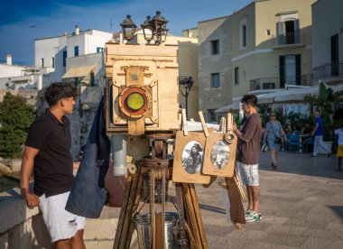 Turistler, antik Otranto, Apulia, İtalya 'nın göbeğinde renkli elbiseler satan bir hediyelik eşya dükkanının önünden geçen kaldırımlı bir sokakta yürürler.
