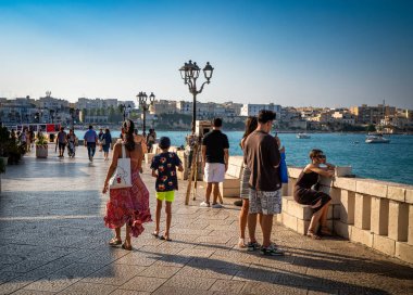 Turistler, antik Otranto, Apulia, İtalya 'nın göbeğinde renkli elbiseler satan bir hediyelik eşya dükkanının önünden geçen kaldırımlı bir sokakta yürürler.