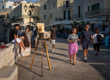 Turistler, antik Otranto, Apulia, İtalya 'nın göbeğinde renkli elbiseler satan bir hediyelik eşya dükkanının önünden geçen kaldırımlı bir sokakta yürürler.