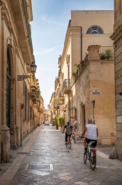 İtalya 'nın antik Lecce kentindeki taş binalarla dolu dar bir sokakta bisiklet süren iki kişinin arka manzarası..