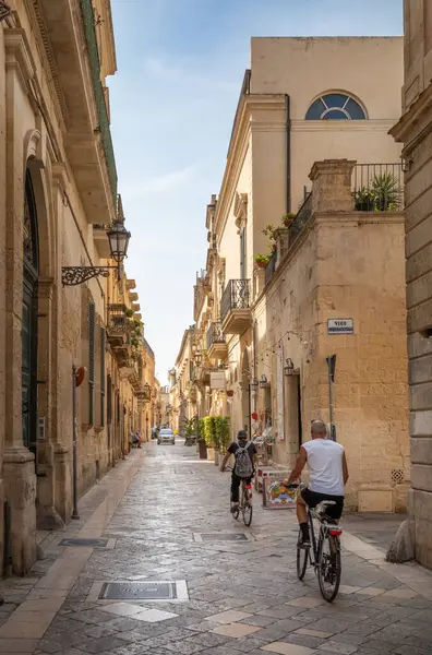İtalya 'nın antik Lecce kentindeki taş binalarla dolu dar bir sokakta bisiklet süren iki kişinin arka manzarası..